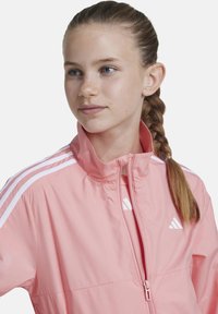 Chaqueta atlética rosa con cuello alto, cremallera frontal y diseño de tres rayas blancas en las mangas. Confeccionada en un tejido ligero y suave.