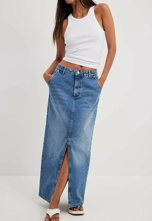 Femme portant un crop top blanc sans manches, une longue jupe en jean bleue avec une fente devant, les mains dans les poches et des sandales blanches à bouts ouverts.