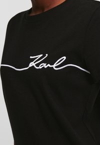 Černé bavlněné tričko s kulatým výstřihem a krátkými rukávy; na hrudi má bílé vyšívané písmo "Karl".