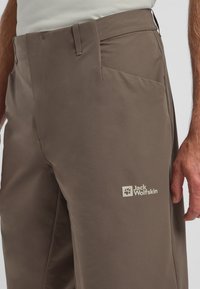 Pantaloni da esterno marroni con una texture liscia, dotati di tasca laterale e logo Jack Wolfskin di un color beige chiaro sulla parte bassa della gamba.
