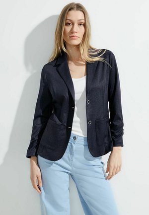 Blazer navy en tissu léger avec col à revers, deux poches avant et boutons argentés, associé à un pantalon bleu clair.