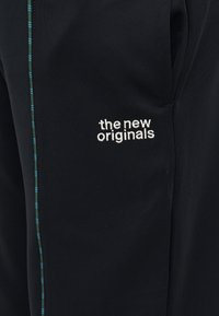 Melns audums ar baltu izšūtu tekstu "the new originals" un šaura vertikāla josla zaļā un zilā pleda rakstā.