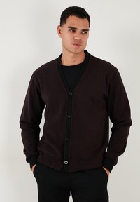 Cardigan marrone scuro con scollo a V, chiusura a bottoni e rifiniture nere a contrasto. Realizzato in tessuto morbido, presenta maniche lunghe e polsini aderenti.