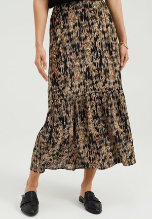 Nainen, jolla on keskipitkä, kerroksittain laskeutuva hame mustalla, ruskealla ja beigen värisellä abstraktilla kuviolla, yhdistettynä mustiin slip-on-kenkiin ja kultaan rannekoruun.