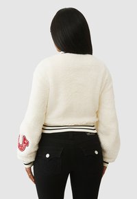 Biała bluza bomber z polaru z czarnymi ściągaczami na mankietach i u dołu, z czerwonym haftowanym logo na lewym rękawie; noszona na czarnych dżinsach.