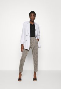 Blazer branco de botão duplo sobre uma polo preta, combinado com calças de xadrez bege e sandálias slide pretas, contra um fundo simples.