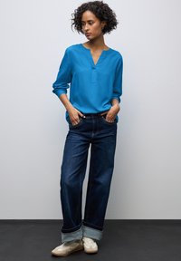 Blauwe blouse met lange mouwen en een V-hals, gecombineerd met een wijde, donkere denim spijkerbroek met opgerolde zomen en witte sneakers.