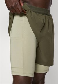Herre shorts med et lett, olivengrønt ytterlag og en tettsittende, lys beige underdel. Begge har myke teksturer og elastiske midjebånd.