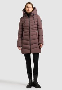 Mujer vestida con un abrigo de invierno acolchado en color malva con capucha, leggings negros y botas negras hasta el tobillo, de pie contra un fondo gris claro.