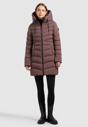 Vrouw in een mauve gewatteerde winterjas met capuchon, zwarte leggings en zwarte enkellaarzen, staand tegen een lichtgrijze achtergrond.