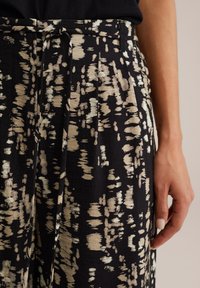 Pantalon en tissu noir avec un motif abstrait beige texturé, taille haute et plis subtils. Détail de ceinture en tissu noué inclus.