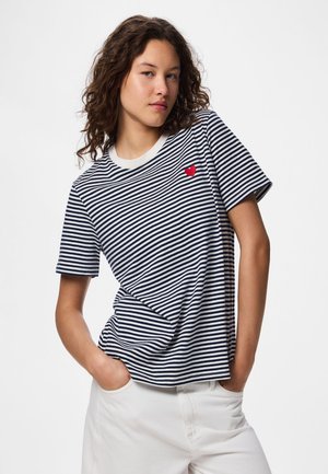 Ung kvinde iført en marineblå og hvidstribet T-shirt med et lille rødt hjerte på brystet, kombineret med hvide bukser, hænder i lommerne.