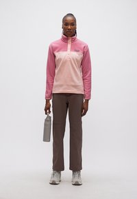 Γυναίκα όρθια, φοράει ροζ φλις μπλούζα, καφέ παντελόνι, λευκά αθλητικά παπούτσια, κρατώντας γκρι μπουκάλι νερού δίπλα της, μπροστά σε απλό φόντο.