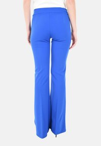 Donna che indossa pantaloni a palazzo, a vita alta, di un blu brillante, in piedi con le braccia rilassate ai lati su uno sfondo bianco semplice.