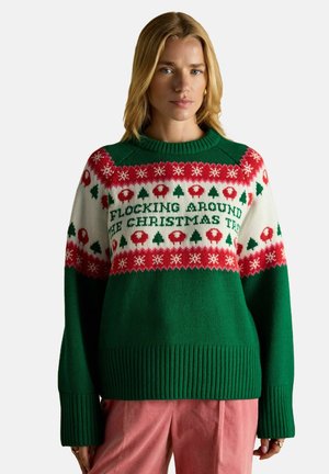 Blonde Frau trägt einen grünen, roten und weißen Weihnachtspullover mit Schafen, Bäumen und dem Text "Flocking Around The Christmas Tree".