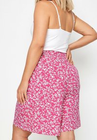 Woman wearing a white sleeveless top and pink floral patterned skirt, shown from the back with one hand on her hip and the other resting by her side.