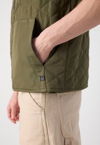 Olive green quilted vest with a side pocket; fabric appears smooth with a slight sheen. Light beige pants with visible stitching detail.
