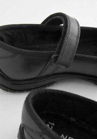 Chaussure en cuir noir avec un bout arrondi, une sangle sur le cou-de-pied, une doublure texturée et une semelle en caoutchouc avec un motif de traction.