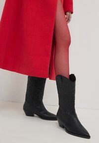 Bottes de cow-boy en cuir noir avec des pointes effilées, des motifs floraux embossés et des talons bas empilés, associées à un manteau rouge.