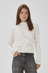 Jeune femme aux longs cheveux bruns portant un haut blanc à manches longues avec un détail drapé et un jean gris foncé avec une ceinture noire.