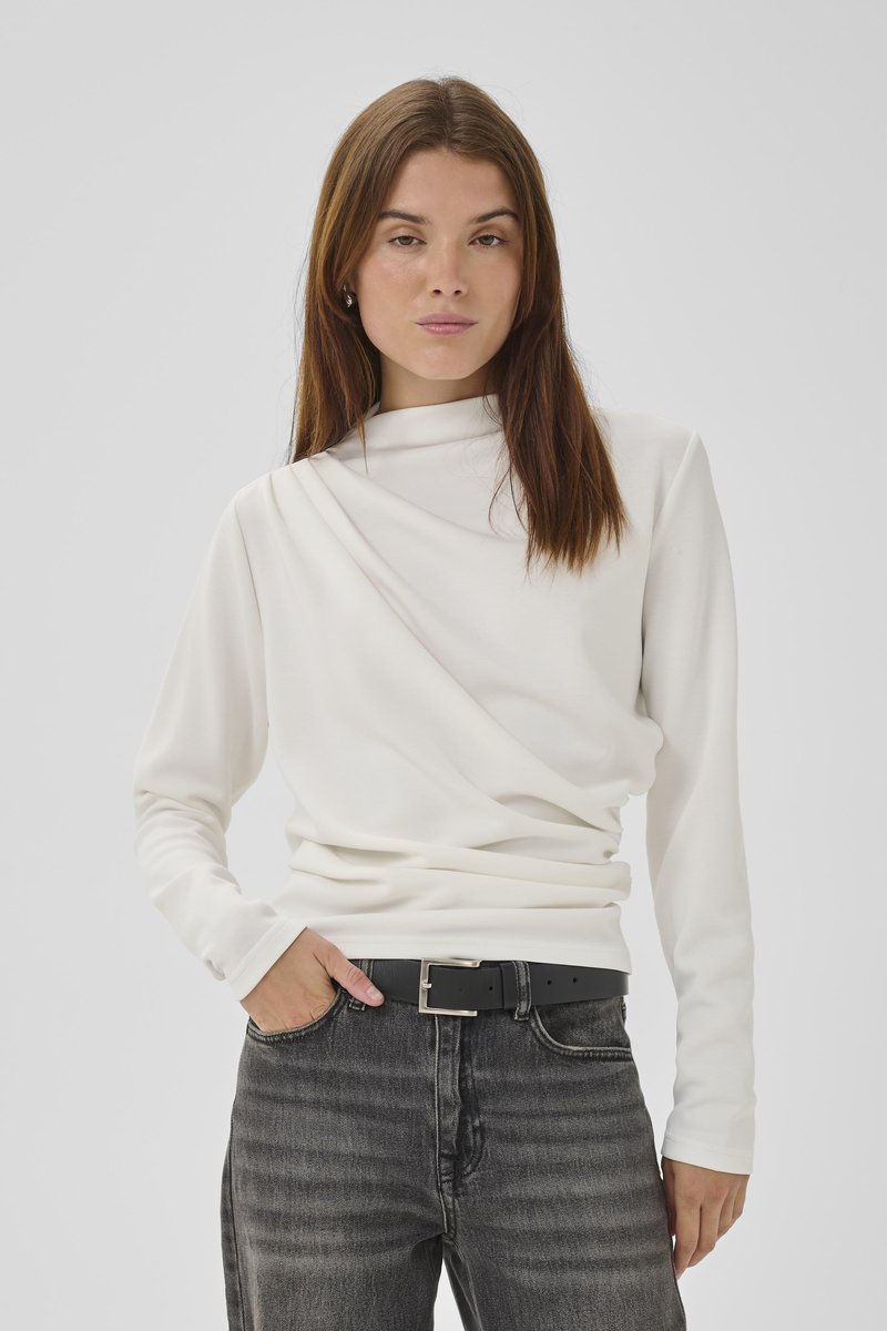 Jeune femme aux longs cheveux bruns portant un haut blanc à manches longues avec un détail drapé et un jean gris foncé avec une ceinture noire.