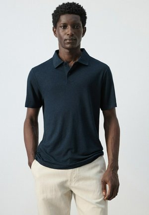 Uomo in piedi con le mani nelle tasche, indossa una polo a maniche corte blu navy e pantaloni beige chiaro su sfondo neutro.