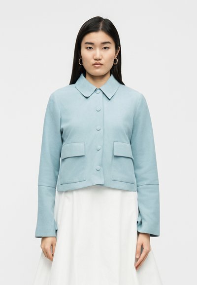 Νεαρή γυναίκα με μαύρα μαλλιά φοράει ένα ανοιχτό μπλε σακάκι με κουμπιά και μπροστινές τσέπες πάνω από μια άσπρη φούστα, στέκεται μπροστά σε απλό φόντο.