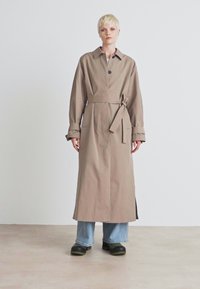 Beige esernyőkabát övvel a deréknál, gombos zárással és hosszú ujjakkal. Világoskék farmerek felett viselve, vastag fekete cipőkkel. Minimalista dizájn.