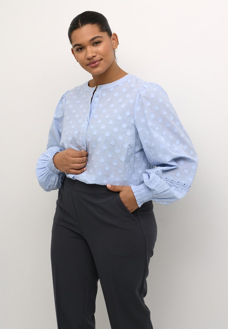 Himmelblaue Bluse mit strukturiertem Punktmuster, Rundhalsausschnitt und Ballonärmeln. Kombiniert mit dunkelgrauen Hosen, die geraffte Bündchen haben.