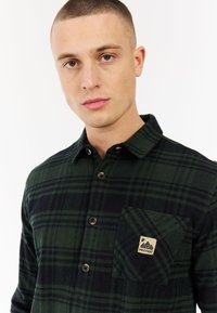 Groene en zwarte geruite flanellen shirt met een knoopsluiting, borstzak en een merkembleem. Zachte textuur en klassieke kraag.
