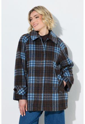 Lächelnde Frau mit blondem Haar, die einen braun-blauen karierten Mantel und blaue Jeans trägt, steht vor einem weißen Hintergrund.