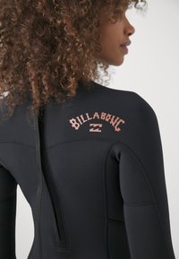 Czarny piankowy kostium z zamkiem na plecach, przeszyciami i wyraźnym różowym logo Billabong na górnej części pleców. Gładka tekstura.