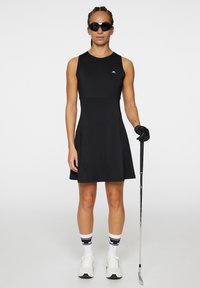 Robe de golf sans manches noire avec un corsage ajusté, une jupe évasée, des poches latérales et un logo de marque ; associée à des chaussures de sport blanches et des lunettes de soleil.