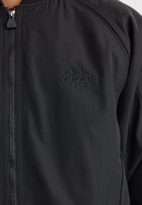 Veste Adidas noire à l'avant avec fermeture éclair et logo Adidas brodé sur la poitrine, portée par une personne.