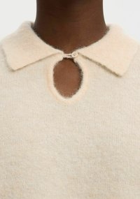 Smetanový svetr s jemnou texturou. Má límeček a klíčový otvor, který je zajištěn kulatým knoflíkem.