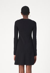 Femme aux cheveux courts portant une robe noire moulante à manches longues jusqu'au genou, vue de dos, avec des boucles d'oreilles dorées sur fond blanc.