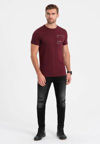Bordeauxrotes T-Shirt mit kurzen Ärmeln, Rundhalsausschnitt und einer Brusttasche. Kombiniert mit schwarzen, verwaschenen Jeans und schwarzen Sneakers.