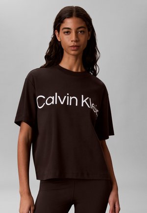 Kvinde med langt mørkt hår iført en løs sort Calvin Klein T-shirt og matchende sorte bukser, vendt fremad mod en ensfarvet grå baggrund.