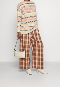 Maglione a maglia oversize con motivi multicolori, abbinato a pantaloni a gamba larga a quadri in arancione e marrone. Una borsa bianca e stivaletti completano il look.