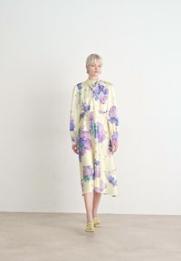 Bloemjurk van gele stof, met paarse en blauwe bloemen, lange mouwen, knopen aan de voorkant en een flowy silhouet, gecombineerd met groene sandalen.