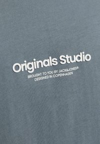 Pelēka T-kreklu audums ar gludu tekstūru, kas rotāts ar reljefa baltiem uzrakstiem: "Originals Studio," "Brought to you by JACK & JONES," "Designed in Copenhagen."