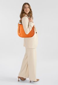 Oranje leren schoudertas met een gestructureerde afwerking, ritssluiting en een gebogen vorm, gedragen over een crèmekleurig pak.