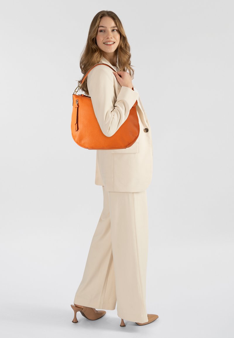 Oranje leren schoudertas met een gestructureerde afwerking, ritssluiting en een gebogen vorm, gedragen over een crèmekleurig pak.
