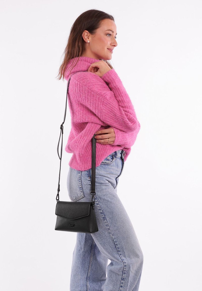 Černá kožená taška přes rameno s klopou, nastavitelným popruhem a strukturovaným povrchem, která je kombinována s růžovým svetrem a světlými džíny.