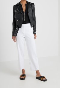 Black leather biker jacket, white tailored cropped pants, and flat black sandals with a textured brown accent. Minimalist design and clean lines.