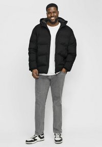Svart pufferjacka med huva, med en slät yta och dragkedjestängning, bärs över en vit t-shirt och grå jeans, kombinerat med svart och vita sneakers.