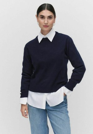 Pull en maille bleu marine avec col rond, porté sur une chemise blanche à col. Les manches sont ajustées et le pull a une texture lisse.