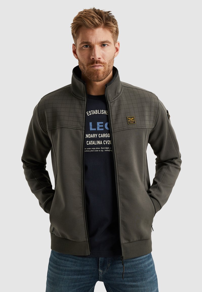 Veste zippée grise avec un motif matelassé sur la poitrine, col montant, poches latérales et poignets côtelés. Portée sur un t-shirt graphique bleu marine.