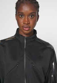 Veste de sport noire avec col montant, dotée de deux poches zippées et de rayures noires brillantes sur les épaules.