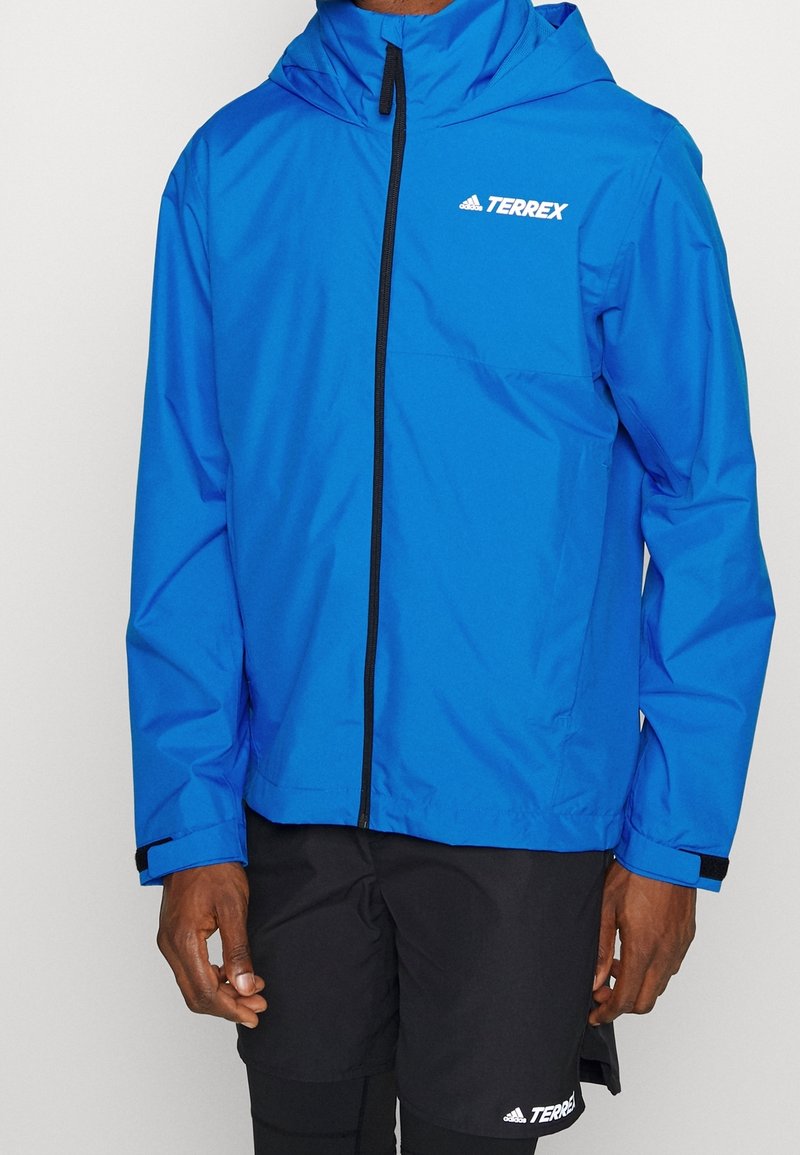 Homme portant une veste à capuche Adidas Terrex bleu vif avec une fermeture éclair frontale et un short noir avec le logo Terrex.